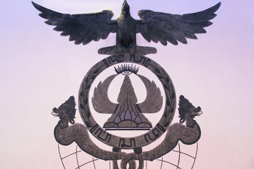 Burung garuda yang tengah mengepakan sayap berdiri di atas lingkaran bertuliskan Purna Wisada, Melambangkan simbol dari Tri Panca Tunggal